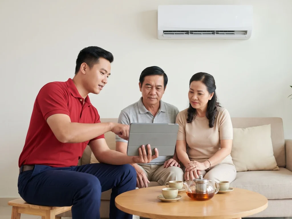 Người thợ áo đỏ dùng máy tính bảng giải thích hạng mục sửa chữa cho hai vợ chồng tại phòng khách